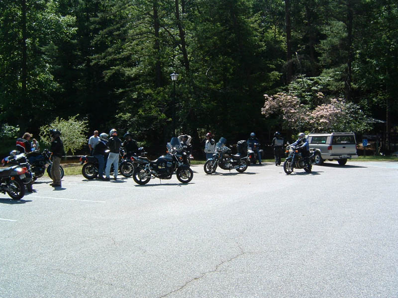 Several motorcycles and people gathered in a paved lot with trees and a van in the background