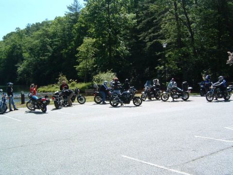 1985 Ford LTD Crown Victoria - Several motorcycles and riders are gathered in a parking lot near a wooded area and a body of water.