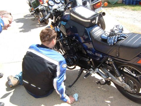 1985 Ford LTD Crown Victoria - Two people examine a Suzuki motorcycle parked outdoors with other bikes nearby.
