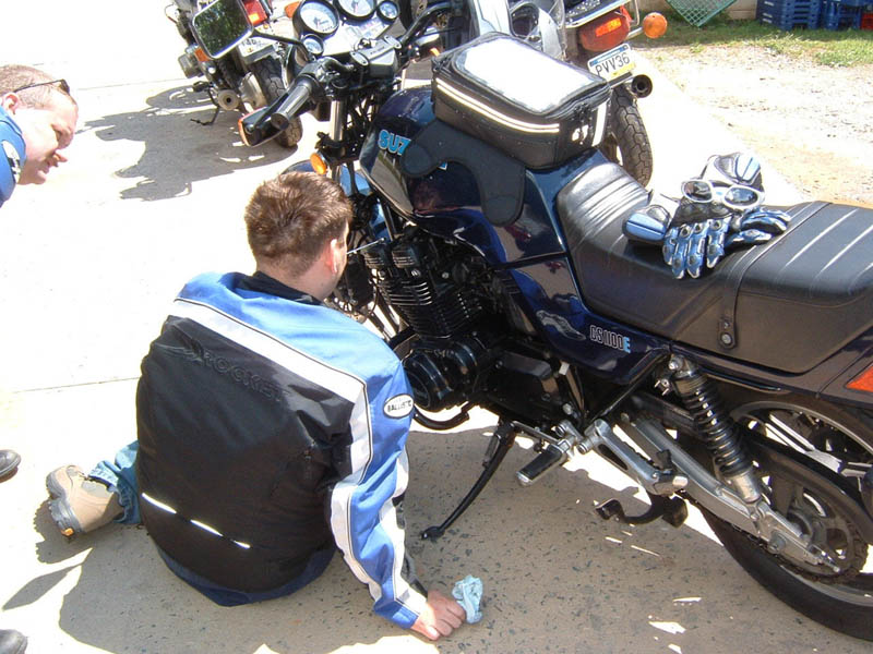 Man in blue jacket looking at a dark blue Suzuki motorcycle. Another motorcycle is behind it