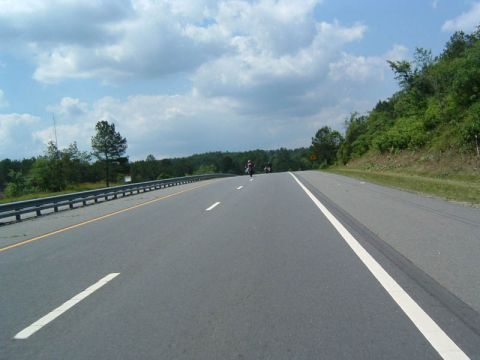 1985 Ford LTD Crown Victoria - Two motorcycles are seen riding on a highway, with trees and a hillside lining the road.