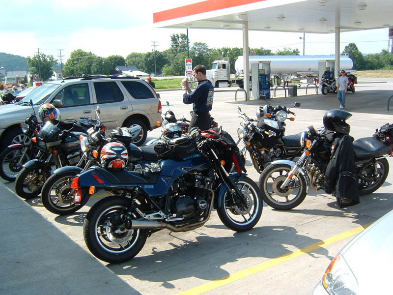 Several motorcycles parked under a gas station canopy. A man is taking a photo with his phone