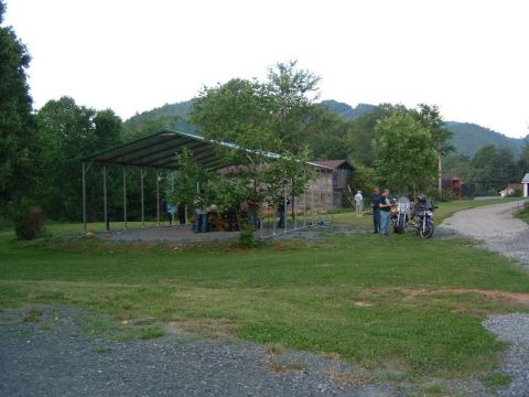 1985 Ford LTD Crown Victoria - Motorcycles are parked near a gathering of people under a shelter, with a barn and wooded hills in the background.