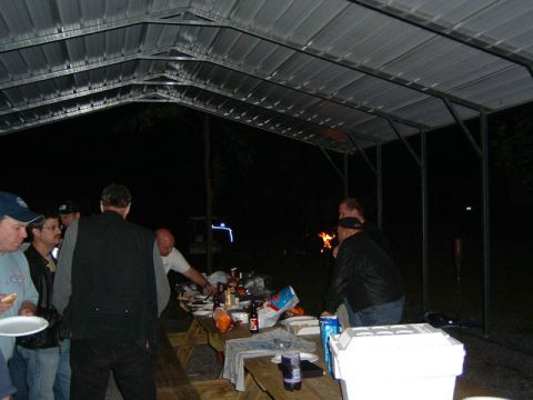 1985 Ford LTD Crown Victoria - A group of people are gathered around a picnic table under a metal canopy at night, with a fire burning in the background.