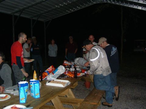 1985 Ford LTD Crown Victoria - A group of people are gathered around a picnic table at night, with food and drinks spread out.