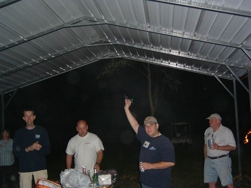 Four men and a woman stand under a metal carport at night, with a table of items in the foreground