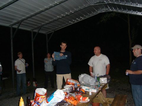 1985 Ford LTD Crown Victoria - A group of people are socializing outdoors at night under a covered shelter, with a picnic table laden with food and drinks.