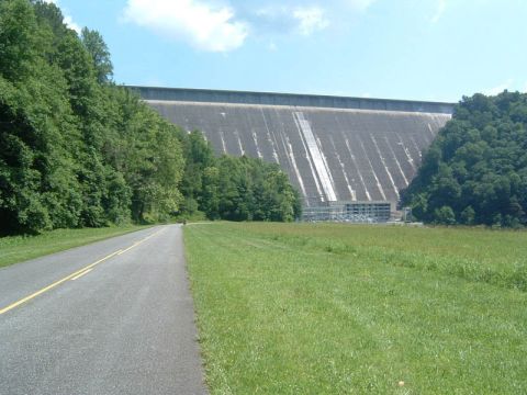 1985 Ford LTD Crown Victoria - A road leads towards a massive concrete dam structure surrounded by trees under a partly cloudy sky.