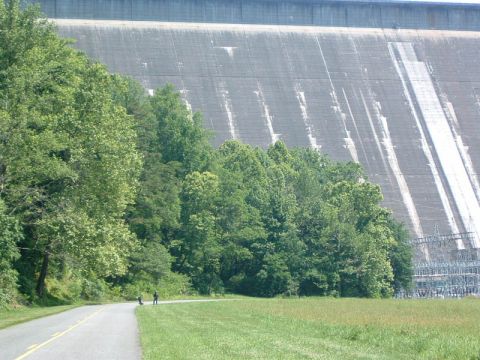 1985 Ford LTD Crown Victoria - A road leads past a field of grass and trees towards a massive concrete dam structure.