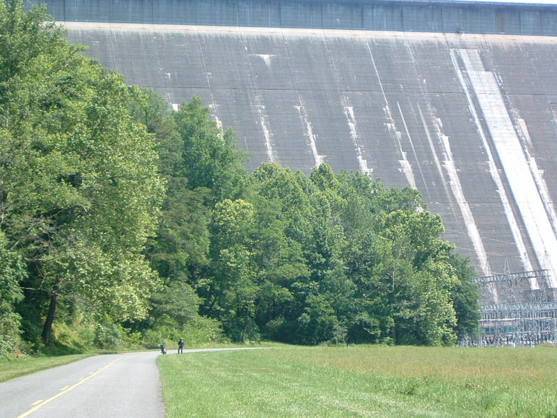 Road curves past grassy field towards a large concrete dam with trees in foreground