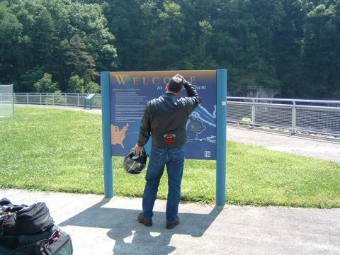 1985 Ford LTD Crown Victoria - A man stands in front of a large sign, reading information about the dam while holding a motorcycle helmet.
