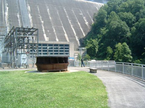 1985 Ford LTD Crown Victoria - A person walks along a paved path next to a large dam and power station complex.