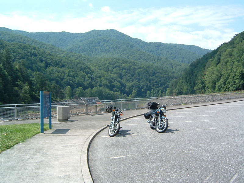 Two parked motorcycles in a paved lot with a forested mountain backdrop