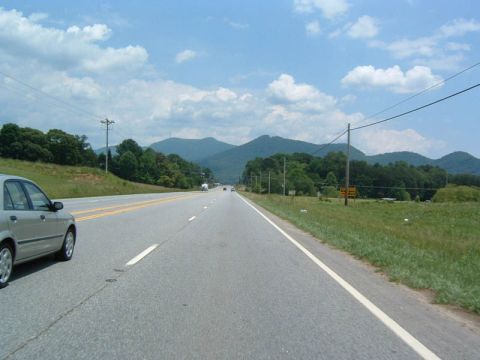 1985 Ford LTD Crown Victoria - A silver sedan travels down a highway with rolling hills and mountains under a partly cloudy sky.