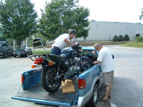 1985 Ford LTD Crown Victoria - Two men load a motorcycle into the bed of a blue pickup truck.