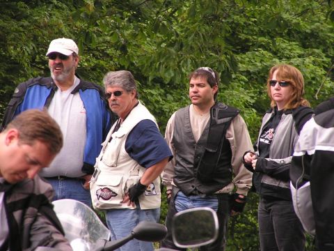 1985 Ford LTD Crown Victoria - A group of motorcyclists pause during a ride, with a motorcycle visible in the foreground and trees in the background.