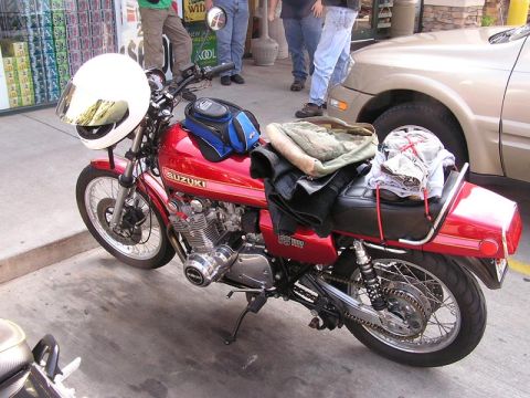 1985 Ford LTD Crown Victoria - A red Suzuki motorcycle is parked curbside, loaded with gear and a helmet.