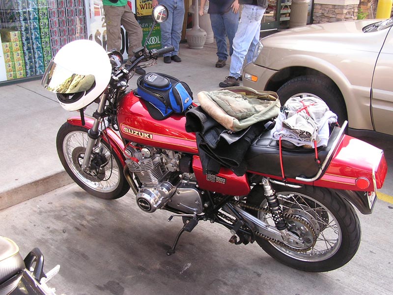 Red Suzuki motorcycle parked next to a tan car. It has a white helmet on the handlebars and bags and clothes strapped to the seat