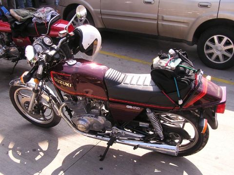 1985 Ford LTD Crown Victoria - A maroon Suzuki motorcycle with a white helmet on its handlebars is parked next to a tan vehicle.