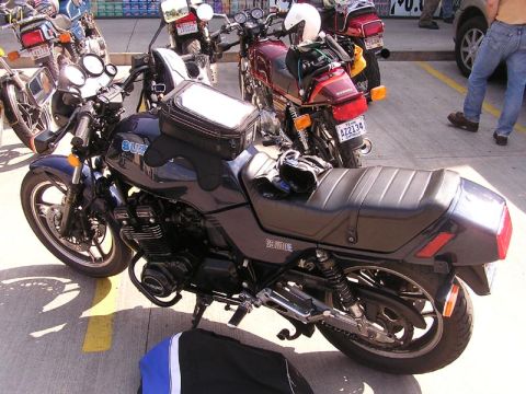1985 Ford LTD Crown Victoria - A dark gray Suzuki GS1000E motorcycle is parked in a lot with other motorcycles visible in the background.