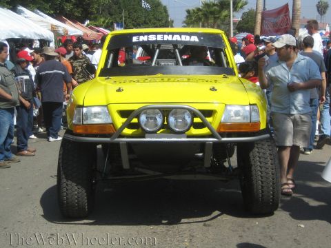 1990 Ford Bronco - yellow off-road race truck with ENSENADA across the windshield and dual driving lights faces a crowd of spectators.