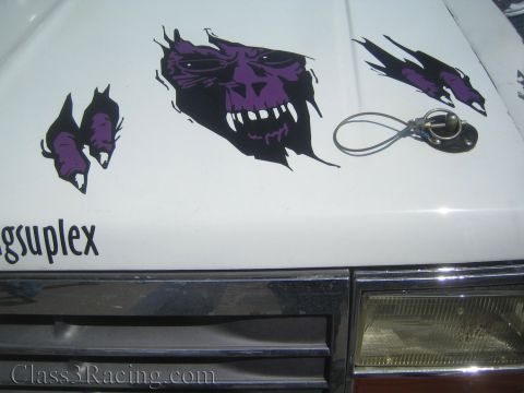 1990 Ford Bronco - close view of a white race truck hood corner shows purple skull graphics, a gsuplex decal and a hood pin above a headlight.