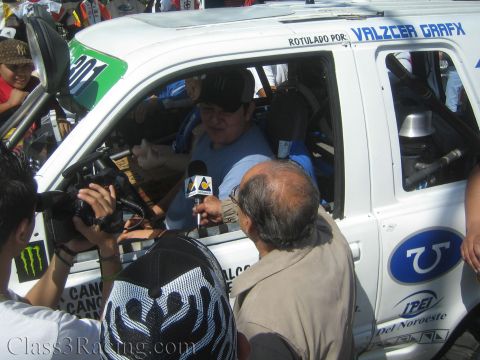 1990 Ford Bronco - capped driver holds a microphone for an interview inside a white race truck lettered Valzcer Grafx as a camera operator films.