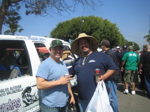 1990 Ford Bronco - Two smiling men, one in a straw hat, pose with canned drinks beside a white Chupakabras race truck in a crowded outdoor venue.