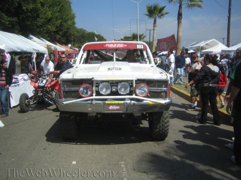 1990 Ford Bronco - Front view of a white off-road race truck numbered 303 with multiple driving lights, parked amid a busy outdoor crowd and tents.