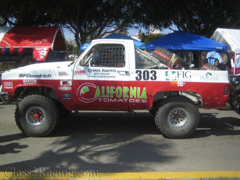 1990 Ford Bronco - Side profile of a red-and-white number 303 'California Tomatoes' off-road race truck parked under canopies at an event.