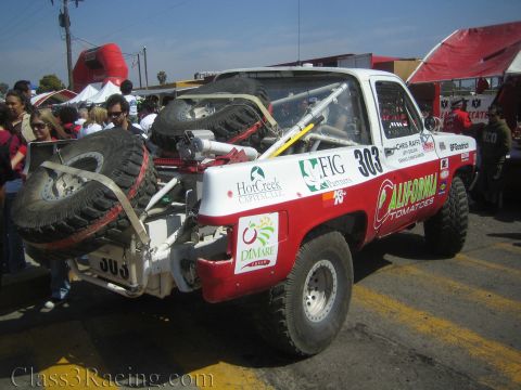1990 Ford Bronco - Rear quarter of a red-and-white number 303 'California Tomatoes' race truck with spare tires on a rack in a crowded race paddock.