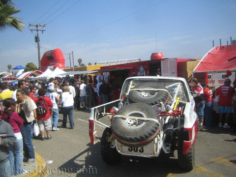 1990 Ford Bronco - Rear view of a white number 303 off-road race truck with mounted spares moving through a crowd near vendor booths.