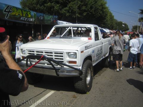 1990 Ford Bronco - white Ford off-road race truck numbered 861 with a roll cage and tube front bumper, surrounded by spectators on a sunny street.