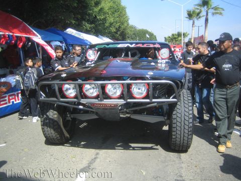 1990 Ford Bronco - black off-road trophy truck marked 'Ensenada' with a tube bumper and four round lights, surrounded by onlookers in the street.