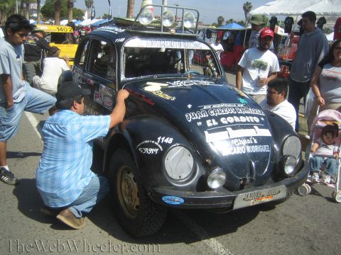 1990 Ford Bronco - black off-road racing Volkswagen Beetle numbered 1148 covered in decals and Spanish memorial text, tended by a man at an event.