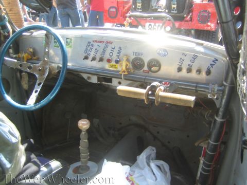1990 Ford Bronco - Interior of a stripped race car showing a blue steering wheel, bare floor, and a metal dash with Spanish-labeled switches and gauges.