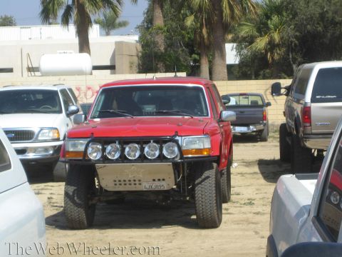 1990 Ford Bronco - red lifted pickup truck with a light bar of round driving lights and big tires sits among other trucks in a dirt parking lot.