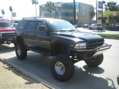 1990 Ford Bronco - black lifted Dodge Durango R/T SUV on tall off-road tires and chrome wheels parked at a street curb with buildings behind.