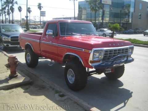 1990 Ford Bronco - red lifted Ford pickup on large off-road tires and chrome wheels sits at a curb beside a fire hydrant on a city street.