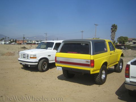 1990 Ford Bronco - yellow lifted Ford Bronco with a hardtop parks on dirt beside a white Ford truck, with houses and power lines in the background.