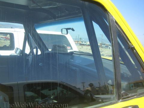 1990 Ford Bronco - Close-up of a yellow vehicle's side window glass reflecting a white truck and open landscape with distant houses.
