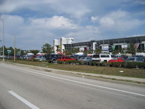 1986 Ford Bronco - A wide view of a parking lot filled with vehicles, with a large stadium structure dominating the background.
