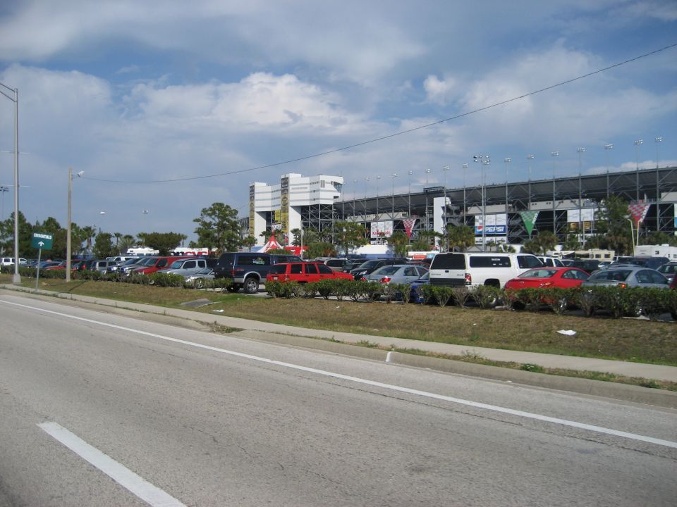 Parking lot with various cars and SUVs, a white van, and a stadium in the background under a cloudy sky