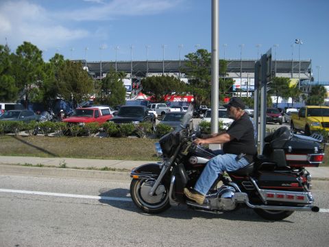 1986 Ford Bronco - A man rides his motorcycle past a parking lot filled with vehicles, with a large stadium visible in the background.
