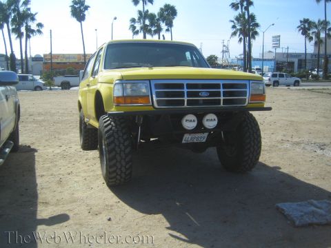 1990 Ford Bronco - yellow lifted Ford SUV fitted with a tube front bumper, dual round driving lights, and large mud-terrain tires sits on a sandy lot under pa…