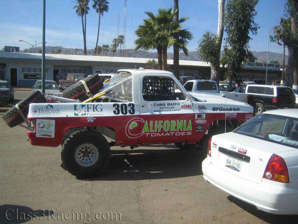 Red and white off-road race truck number 303 with California Tomatoes livery and spare tires parked in a lot