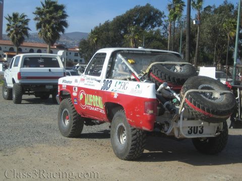 1990 Ford Bronco - rear quarter of a red-and-white number 303 race truck shows bed-mounted spare tires and a roll cage, parked on dirt near other trucks.