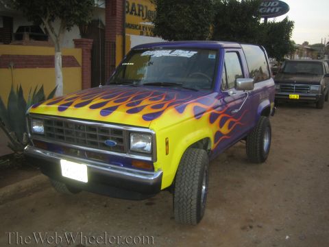 1990 Ford Bronco - two-door SUV painted purple with vivid yellow-and-orange flames sits curbside, its Ford grille and lift kit visible against storefronts.