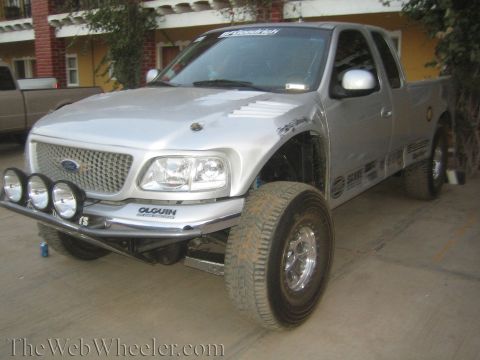 1990 Ford Bronco - silver lifted Ford extended-cab pickup with a prerunner tube bumper and four round driving lights is parked in a tiled courtyard.