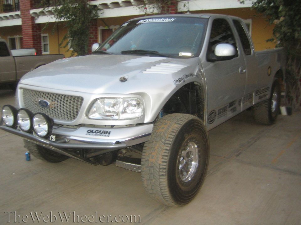 Silver lifted Ford extended-cab pickup with tube bumper and dual driving lights in a courtyard